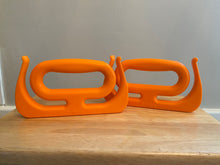 Load image into Gallery viewer, The two orange Mammoth Grips are sitting on a wood table. They have a large handle with two hooks that come up on either side, for bags or other items that need to be carried.
