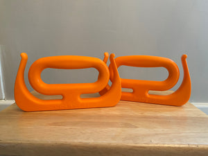 The two orange Mammoth Grips are sitting on a wood table. They have a large handle with two hooks that come up on either side, for bags or other items that need to be carried.