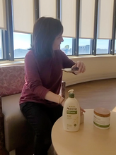 Load image into Gallery viewer, Hannah using her phone to read product labels. A bottle of Aveeno lotion is sitting on a table in front of Hannah, who has brown hair and a red shirt.
