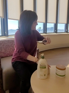 Hannah using her phone to read product labels. A bottle of Aveeno lotion is sitting on a table in front of Hannah, who has brown hair and a red shirt.
