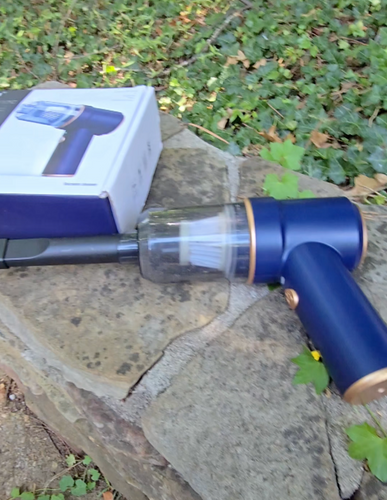 A blue device with gold accents is laying on a stone wall beside a box with its image on it. There is slender extension piece on the front of it. The device is the shape of a handheld blow dryer.