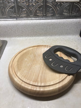 Load image into Gallery viewer, A mezzaluna knife with a black handle is sitting on a wood, circular cutting board. The blade of the knife is covered with a black sheath.
