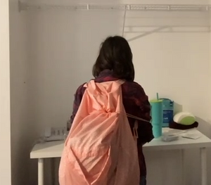 A person with long brown hair is wearing a large neon orange laundry backpack on her back. The drawstring is pulled around to the front.