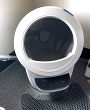 Load image into Gallery viewer, A white round machine is sitting on the floor, with a black step up to a large round opening. On the edge is a litter guard, and inside is cat litter. On one side there is a grate that will comb through the cat litter.
