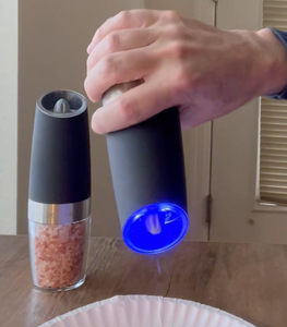 A person is tilting the pepper mill downward, activating the grinder and the blue light, as pepper comes out. The salt grinder, filled with coarse pink salt, is sitting on the table.