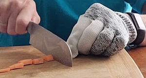 A person is using a large knife to chop a carrot on a cutting board. The hand that is holding the carrot (not holding the knife) has a gray and white fabric (of some type) glove on it. 