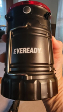 Load image into Gallery viewer, Side of the black body of the camping lantern showing a metal handle that&#39;s folded down, a black button about the size of a thumb for the flashlight, and &quot;eveready&quot; branded in the middle.
