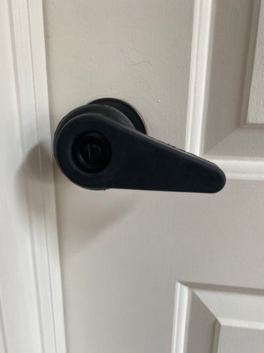 Sammons Preston black rubber extension handle creates a lever on a black round doorknob, on a white door. The key hole is hard to see because it is also black, but there is an opening in the handle for the keyhole.