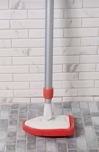 The white scrubber head has a red sponge attached to it, shaped to be able to fit into corners. It has a silver metal pole. Standing straight up on a gray tiled floor with a white tiled wall behind it.
