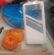 Load image into Gallery viewer, Surface of the mandoline slicer. The top part is clear, the middle blade is gray, and the bottom is white. The frame is white. A tomato and arm of a person are visible behind the mandoline slicer. 
