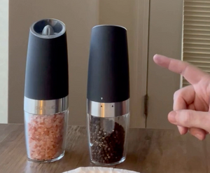 A person is pointing at the gravity electric salt and pepper grinder set. Each device has a large black top, where the grinder is, and a clear base, through which you can see the spices.