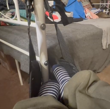 Load image into Gallery viewer, The black Travel foot rest is hanging from the metal pole of a bed and a person wearing striped socks has their feet up using it.
