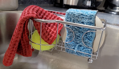 Simple Houseware silver kitchen sink sponge holder with a blue dish sponge, a yellow round sponge, a white drain cover, and a red dish cloth hanging over the edge. 