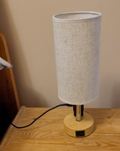 Load image into Gallery viewer, Small bedside table touch lamp with three charging ports on the base. It has a tall round off-white lamp shade and a wooden base with a metal lamp post.
