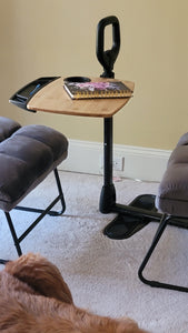 Bamboo tray table with a black tray attachment, a black cup holder, and a black stand assist handle positioned between a gray chair and a foot rest.