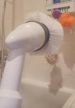 Load image into Gallery viewer, White round brush head with gray accent. Behind it is a bathtub, white tile shower wall, and a shower caddy with an orange loofah and other shower products.
