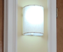 Load image into Gallery viewer, A rounded white wall sconce hanging on a yellow wall between two door frames is lit up.
