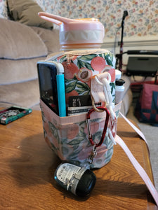 Peach patterned Cube Water Bottle sitting on a table holding a phone, credit card and money, and lotion in its pockets, as well as hand sanitizer clipped to it.
