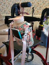 Load image into Gallery viewer, Peach patterned Cube Water Bottle hanging from the handle of a rollator and holding a phone and lotion in its pockets, as well as keys clipped to it.
