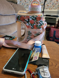 The contents that the mesh pocket of the water bottle normally holds is laying on a table in front of it. There is a phone, credit card, keys, lotion, sanitizer, and medicine. The long strop of the water bottle is visible, as is the large handle of the bottle.