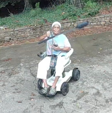 Load image into Gallery viewer, Karen holding the ZoomBroom up on the air with one hand. She is seated in a white power chair.
