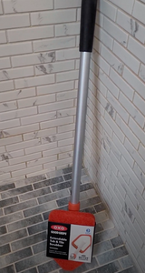 OXO Tub & Tile Scrubber leaning upright in the corner of a tile shower. It has a black grip as the handle, a silver extendable arm, and a red sponge cleaning head shaped like an arrow for corners. The packaging is still on around the scrubber and says "OXO Good Grips Extendable Tub & Tile Scrubber."