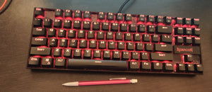 Black keyboard with red backlighting. A pencil is in front of the keyboard and is approximately one third of the keyboard's length. 
