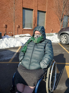 Ericka bundled up in her dark green (called "darkest spruce" by the manufacturer) down jacket with a scarf wrapped around her face. She is in her wheelchair in a parking lot. There is snow in the background. 