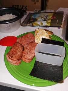 The clear mold is sitting on a piece of seaweed on a green plate  beside several pieces of spam and fried eggs.