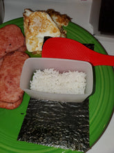 Load image into Gallery viewer, Rice is in the clear mold, sitting on top of a piece of seaweed with a small red spatula beside the mold. All of this is on a green plate along with spam and fried eggs.
