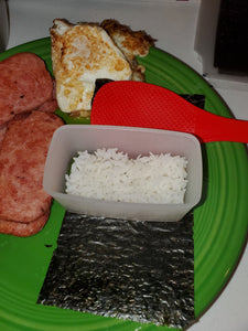 Rice is in the clear mold, sitting on top of a piece of seaweed with a small red spatula beside the mold. All of this is on a green plate along with spam and fried eggs.