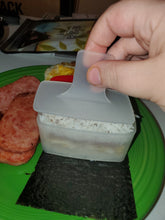 Load image into Gallery viewer, Musubi mold in action, with all the ingredients in the mold and a person holding the plunger, about to put it in. There is also spam and fried eggs visible on the plate.
