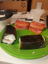 Load image into Gallery viewer, The finished musubi! Two finished musubi are wrapped in seaweed, with rice and other ingredients visible in the middle. There is extra spam on the green plate.
