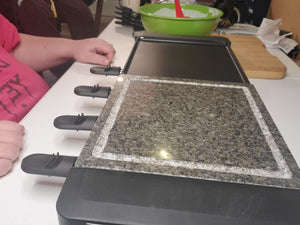The raclette grill is sitting on a table with a green bowl beside it. A person is sitting at the table, but only a pink shirt and their hands are visible.