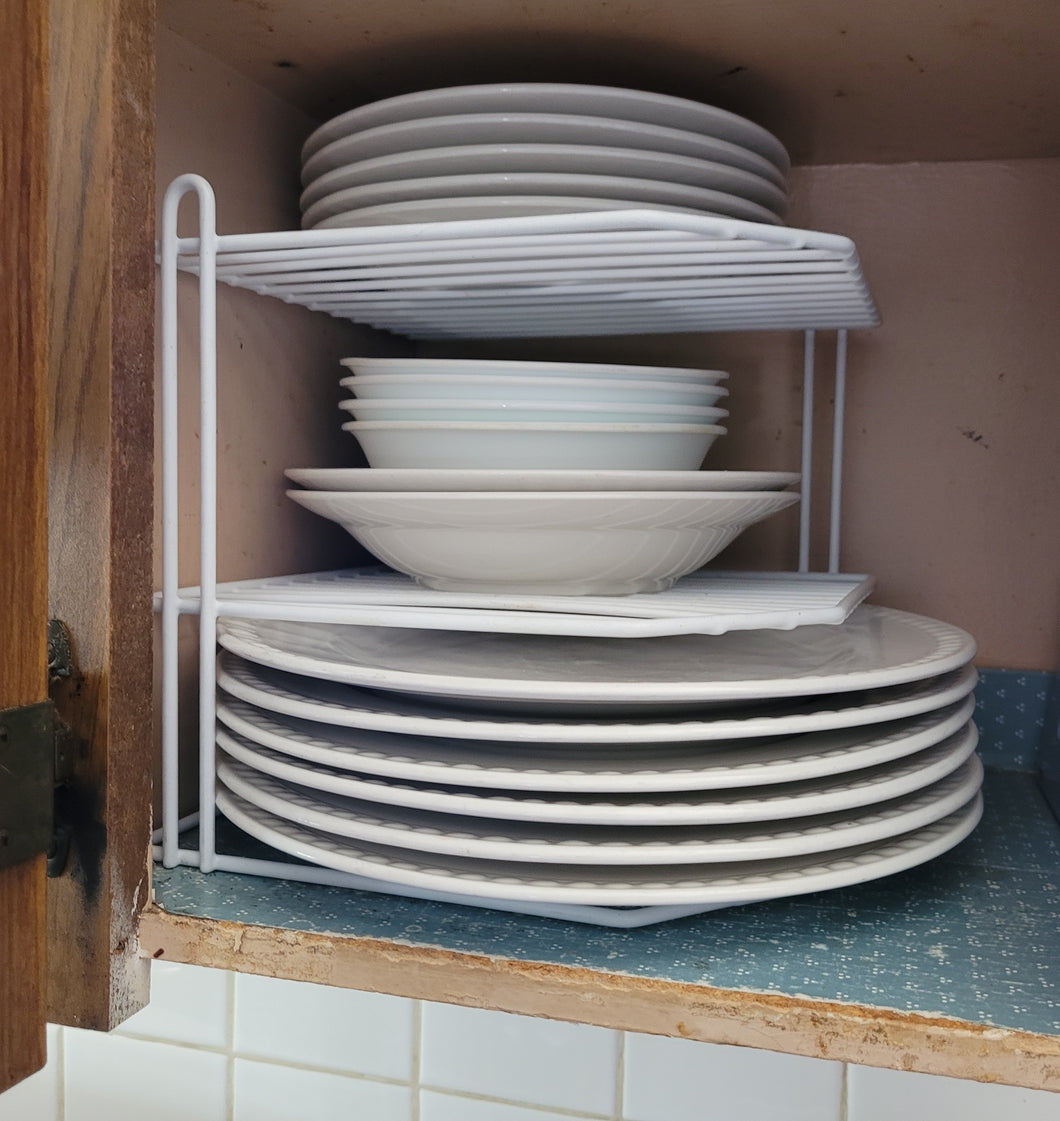 White corner rack sitting in a wooden kitchen shelf. On the bottom (sitting directly on the cabinet) are six large kitchen plates. On the second shelf are two large bowls and four smaller bowls. On the top shelf are five smaller plates.