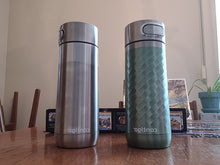 Load image into Gallery viewer, Two Contigo Luxe Autoseal mugs standing side by side. One is silver and the other has a silver lid and a green base with a geometric pattern in it.
