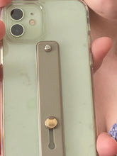 Load image into Gallery viewer, The olive green strap in the flat position, secured to the back of a phone. A silver button at the top is visible, and a slightly larger shiny button appears at the bottom, where space in the strap allows it to be pulled up or pushed back down to lay flat.
