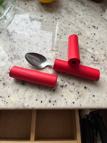 A spoon with a red foam grip is sitting on a countertop alongside to other red foam grips.