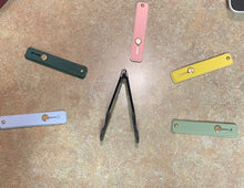 Load image into Gallery viewer, Five of the phone straps are laid out on the floor, all in different colors including light blue, dark green, pink, yellow, and light green. The free kickstand is black and sits in the middle.
