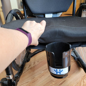 A person's hand is lifting the cushion to show that the HandiCup is held in place by a plastic extension that slips underneath the wheelchair seat cushion. 