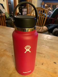 32 oz. wide mouth Hydro Flask water bottle in the color "snapper", which appears to be a bright reddish-pink. The hydro flask logo is in the middle of the bottle, with "Hydro Flask" written at the bottom. The lid is black and has a handle on it for carrying that is in the upright position above the bottle.