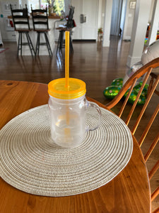 Plastic mason jar cup with handle sits on a placement on a table. The cup is clear with a neon orange lid and straw.