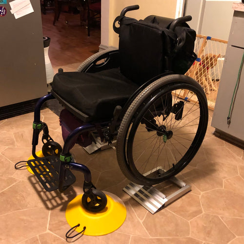 A black wheelchair in a kitchen has its front wheels sitting on yellow wheel docks and the back wheels sitting in the rolling wheel stands by Maxxhaul.