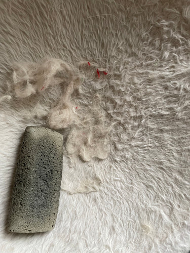 Fur-Zoff pet hair removal tool - which is dark gray and looks like a large, rough stone - is sitting on a white furry cat hammock with visible white pet hair around it. 