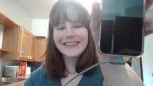 Load image into Gallery viewer, White woman with brown hair and bangs smiling and holding up a phone with the gray 90 degree charging cable plugged into the bottom of it. The cable extends to the side rather than straight down.
