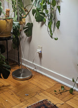 Load image into Gallery viewer, A Kasa Smart Plug is plugged into an outlet. There is a lamp plugged into the smart plug. A blue light is glowing on the side. A silver floor lamp sits close to the outlet, and there are plants all around.
