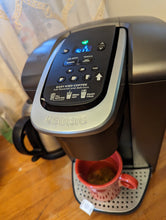 Load image into Gallery viewer, Top view of Keurig machine (black with silver accents). Ten small buttons and a small screen appear on top. A silver handle says Keurig on it. A red mug is underneath the machine with a tea bag in it. 
