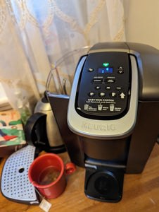 View of Keurig machine showing the lid off the water tank (on the side of the machine) and the drip tray pulled away, making it possible to put larger cups or bottles underneath the machine. 