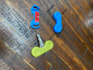 Silver key with the yellow Keywing Key Turner on it, beside a blue keywing key turner without a key, and the inside of a key turner (showing a 3M sticky strip).