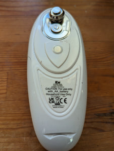 Bottom of the Kitchen Mama can opener, white and showing the stainless steel parts that cut open the cans. Text reads "Caution for use only with AA battery. Household use only"